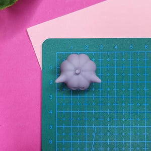 May include: A light purple, 3D printed, pumpkin-shaped object with a stem on top. The object is sitting on a green cutting mat with a grid pattern.