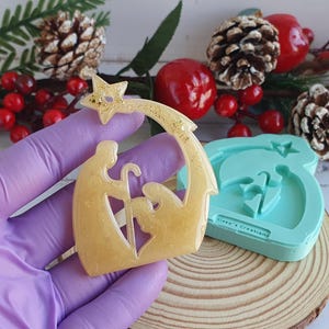 Op de afbeelding: Een gouden hars ornament dat de kerststal voorstelt met een ster en een kribbe. Het ornament wordt in een handschoen gehanteerd en een blauwe siliconen mal van het ornament is zichtbaar op de achtergrond.
