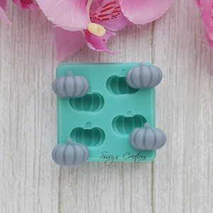 May include: A teal silicone mold with five gray pumpkin-shaped soap embeds. The mold has the text "Sissy's Creations" in a cursive font. The background includes pink flower petals.