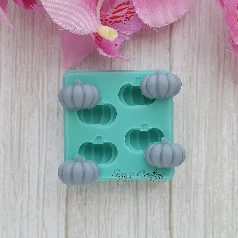 May include: A teal silicone mold with five gray pumpkin-shaped soap embeds. The mold has the text "Sissy's Creations" in a cursive font. The background includes pink flower petals.