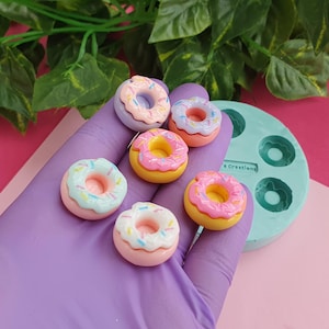 Puede incluir: Seis mini donuts con coloridos espolvoreos y glaseado en una variedad de colores, incluyendo rosa, amarillo y azul. Las donas están dispuestas en una mano enguantada de morado y un molde de silicona verde.