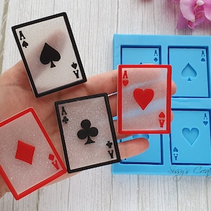 Puede incluir: Cuatro cartas de as de juego de resina transparente con diferentes palos: picas, corazones, diamantes y tréboles. Las cartas están encima de un molde de silicona azul con cuatro formas de cartas vacías.