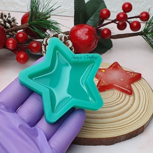 Puede incluir: Un molde de silicona verde con forma de estrella. El molde se sostiene en una mano enguantada. Una pieza de resina roja en forma de estrella es visible en el fondo.