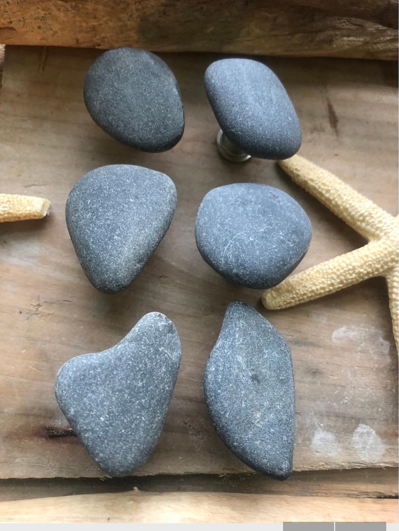 May include: Six gray stone cabinet knobs with a natural, organic look. The knobs are various shapes and sizes, and are arranged on a wooden surface with a starfish. These knobs would be a unique addition to any home decor.