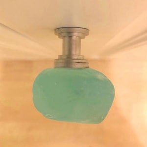 Op de afbeelding: Een lichtblauwe glazen knop met een zilveren metalen basis. De knop is bevestigd aan een wit plafond.