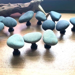 May include: A collection of gray stone cabinet knobs with black stems, arranged on a wooden surface. The knobs have a natural, organic shape, and are accompanied by a piece of driftwood.
