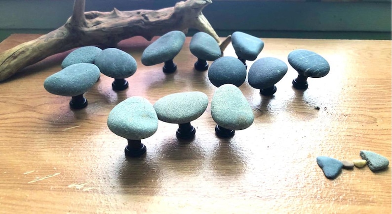 May include: A collection of gray stone cabinet knobs with black stems, arranged on a wooden surface. The knobs have a natural, organic shape, and are accompanied by a piece of driftwood.