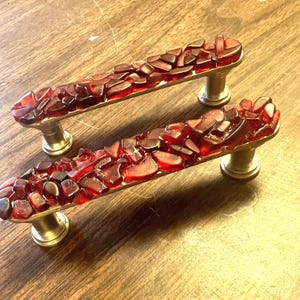May include: Two cabinet handles with a rectangular design. They feature a brushed metal base and are adorned with a mosaic of red glass fragments. The cabinet hardware is designed for installation on a cabinet or drawer, adding a decorative touch.
