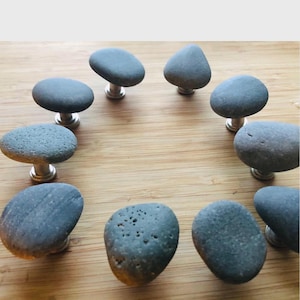Może przedstawiać: Kolekcja dziesięciu szarych kamiennych gałek do szafek ze srebrnymi trzpieniami. Gałki wykonane są z naturalnych kamieni rzecznych, każda o unikalnym kształcie i fakturze. Ułożone są na jasnobrązowej drewnianej powierzchni.