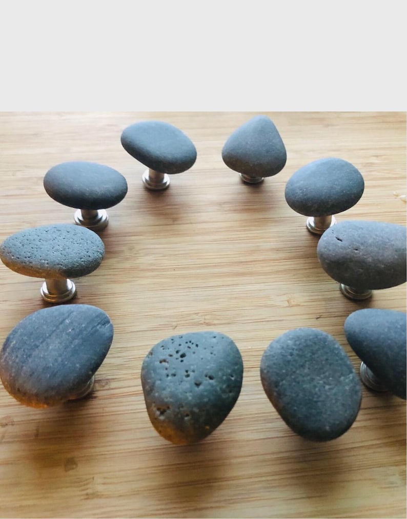 May include: A collection of ten gray stone cabinet knobs with silver-colored stems. The knobs are made from natural river stones, each with a unique shape and texture. They are arranged on a light brown wooden surface.