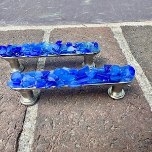 May include: Two cabinet pulls with a brushed nickel finish and a bed of vibrant blue sea glass. The pulls are rectangular with rounded ends and are elevated on decorative feet. The sea glass is a mix of shades of blue.