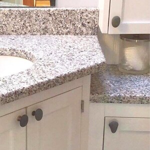 May include: A bathroom vanity with a granite countertop. The countertop has a speckled pattern of gray, white, and brown. White cabinets with dark gray knobs are visible. A glass jar with a candle is on the countertop.