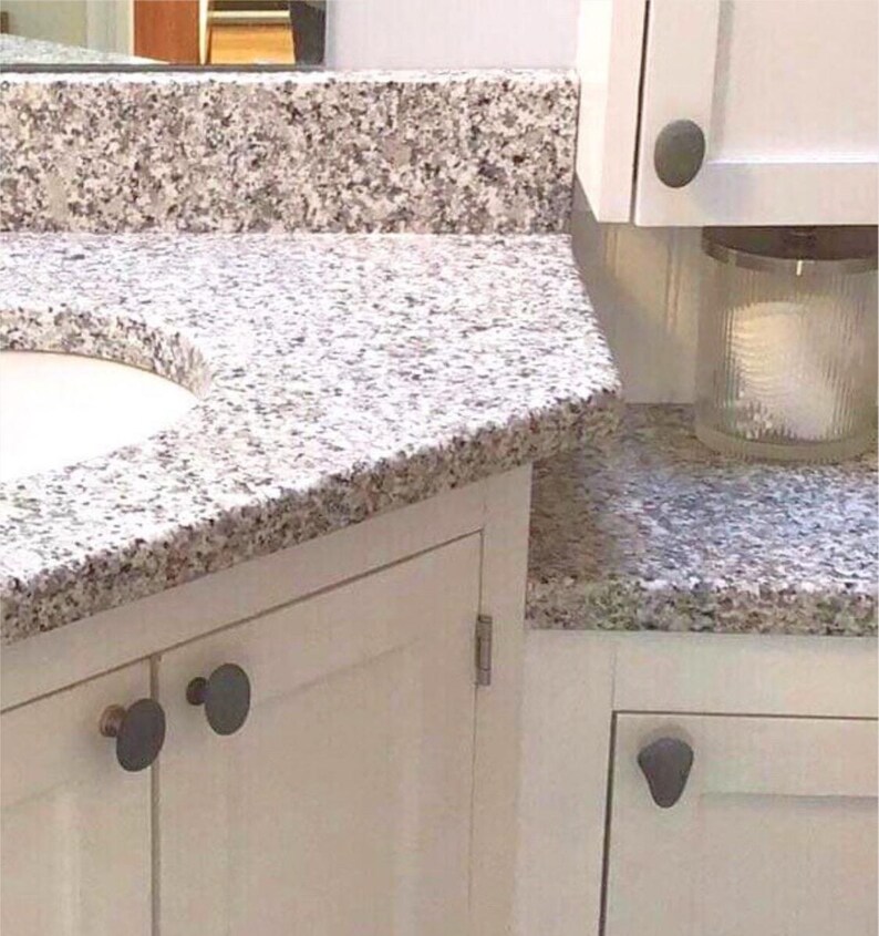 May include: A bathroom vanity with a granite countertop. The countertop has a speckled pattern of gray, white, and brown. White cabinets with dark gray knobs are visible. A glass jar with a candle is on the countertop.