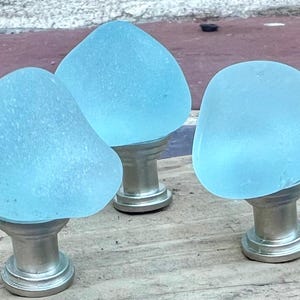 May include: Three light blue sea glass cabinet knobs with silver-coloured metal bases. The knobs are smooth and rounded, with a frosted appearance. They are arranged on a weathered wooden surface.