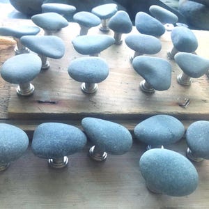 May include: A collection of gray stone cabinet knobs with silver-toned bases. The knobs are made from smooth, rounded stones, each unique in shape. They are arranged on a wooden surface, suggesting a rustic or natural aesthetic. A starfish is visible in the background.