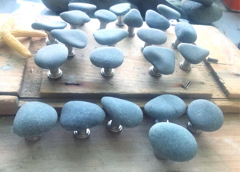 May include: A collection of gray stone cabinet knobs with silver-toned bases. The knobs are made from smooth, rounded stones, each unique in shape. They are arranged on a wooden surface, suggesting a rustic or natural aesthetic. A starfish is visible in the background.