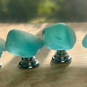 May include: Four cabinet knobs featuring smooth, translucent, light blue sea glass tops with irregular, rounded shapes. Each piece is securely attached to a polished, reflective silver-toned metal base with a circular, tiered design.