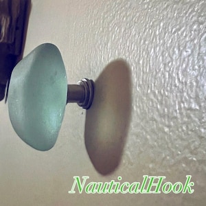 May include: A wall-mounted hook featuring a smooth, rounded, pale seafoam green glass knob attached to a metallic silver-toned base. The text "NauticalHook" is printed in a green font at the bottom right corner of the image.