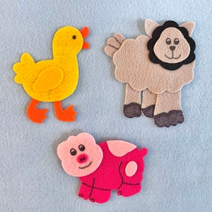 May include: Three felt animal figures: a yellow duck, a gray sheep with a black face, and a pink pig. The figures are on a light blue background.