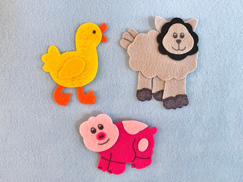 May include: Three felt animal figures: a yellow duck, a gray sheep with a black face, and a pink pig. The figures are on a light blue background.