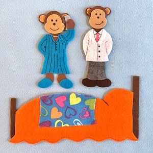 May include: Two felt monkey figures, one in blue pajamas and one in a white coat, stand next to a felt bed with an orange headboard and a blue and pink heart patterned blanket.