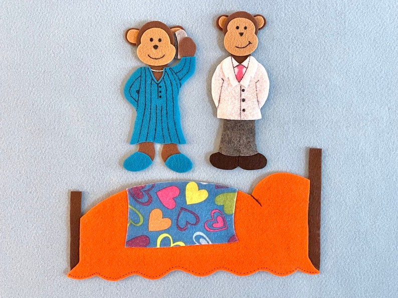 May include: Two felt monkey figures, one in blue pajamas and one in a white coat, stand next to a felt bed with an orange headboard and a blue and pink heart patterned blanket.