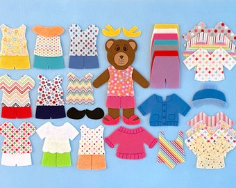 May include: A brown felt bear doll with pink, white, and green clothing. The bear is surrounded by a variety of felt clothing pieces, including shirts, pants, dresses, and a hat. The clothing pieces are all different colors and patterns.
