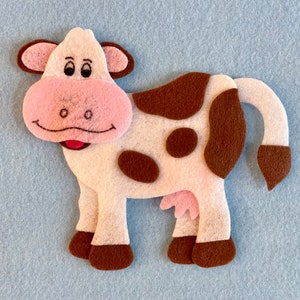 May include: A white felt cow with brown spots, pink ears, and a pink nose. The cow has a brown tail and brown hooves.