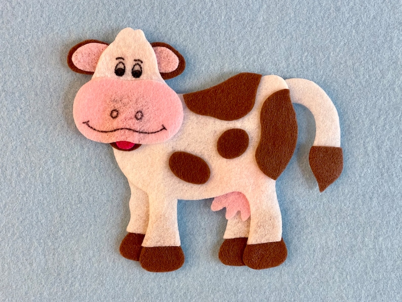 May include: A white felt cow with brown spots, pink ears, and a pink nose. The cow has a brown tail and brown hooves.