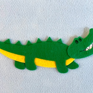 May include: A green felt alligator with yellow felt accents. The alligator has a toothy grin and is lying on a light gray surface.