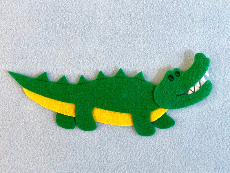 May include: A green felt alligator with yellow felt accents. The alligator has a toothy grin and is lying on a light gray surface.