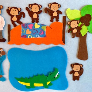 May include: Felt toy set featuring a brown alligator, a blue pond, a brown palm tree with coconuts, and six brown monkeys. One monkey is wearing a blue and white striped shirt, another is wearing a white doctor's coat, and the rest are wearing nothing. The monkeys are on a bed with a red and orange headboard and a blue and green blanket with pink hearts.