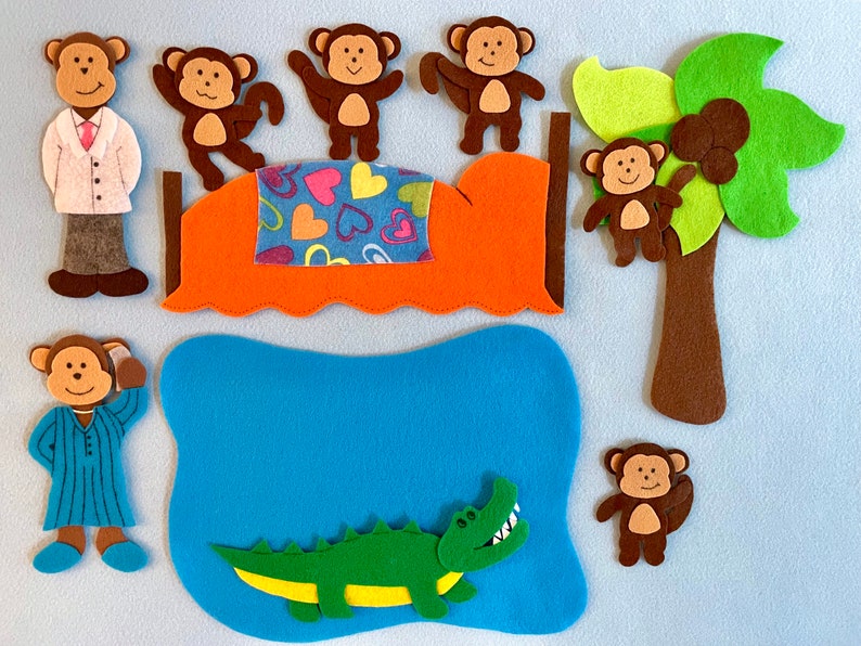 May include: Felt toy set featuring a brown alligator, a blue pond, a brown palm tree with coconuts, and six brown monkeys. One monkey is wearing a blue and white striped shirt, another is wearing a white doctor's coat, and the rest are wearing nothing. The monkeys are on a bed with a red and orange headboard and a blue and green blanket with pink hearts.
