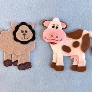 May include: A brown felt sheep with a black felt mane and a white felt cow with brown spots. Both animals have smiling faces.