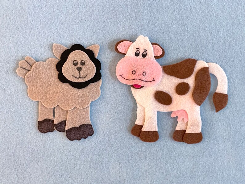 May include: A brown felt sheep with a black felt mane and a white felt cow with brown spots. Both animals have smiling faces.