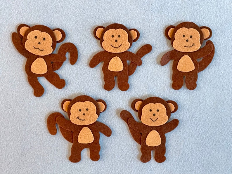 May include: Five brown felt monkey cutouts with smiling faces. The monkeys have brown felt tails and are standing on a light blue surface.