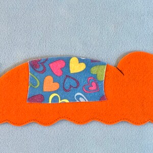 May include: A felt bed with a blue and multi-colored heart patterned blanket. The bed is orange with brown felt side rails.