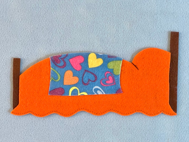May include: A felt bed with a blue and multi-colored heart patterned blanket. The bed is orange with brown felt side rails.