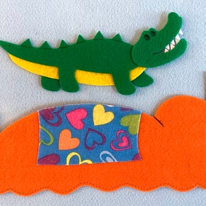 May include: A green felt alligator with yellow accents is jumping over a bed with an orange headboard and a blue and pink heart patterned blanket.