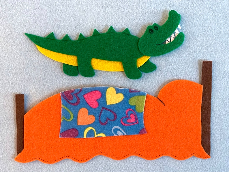 May include: A green felt alligator with yellow accents is jumping over a bed with an orange headboard and a blue and pink heart patterned blanket.