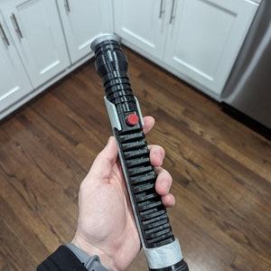 May include: A black and silver lightsaber prop with a red button. The lightsaber is held in a person's hand.