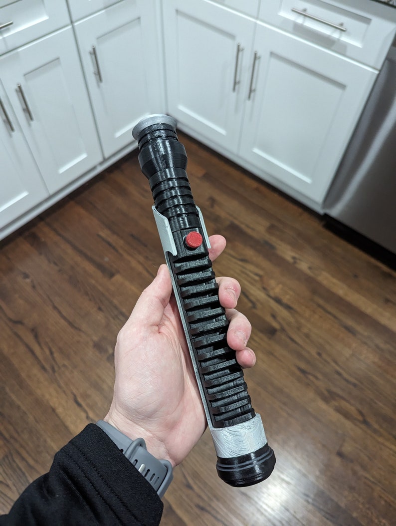 May include: A black and silver lightsaber prop with a red button. The lightsaber is held in a person's hand.