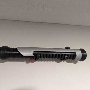 May include: A black and silver lightsaber prop with a red button. The lightsaber is mounted on a wall.