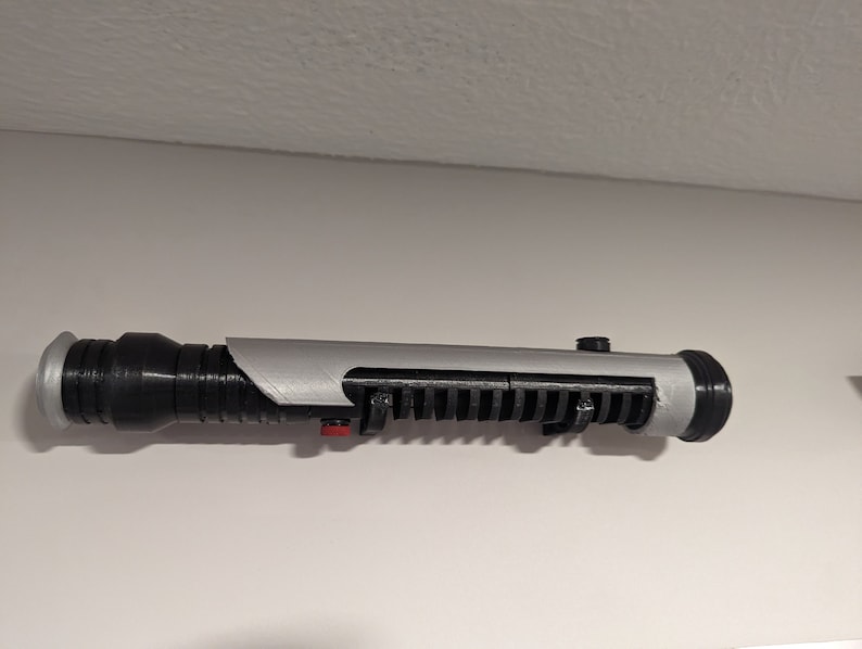 May include: A black and silver lightsaber prop with a red button. The lightsaber is mounted on a wall.