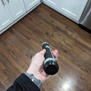 May include: A black and grey lightsaber toy with a red button. The lightsaber is being held in a hand.