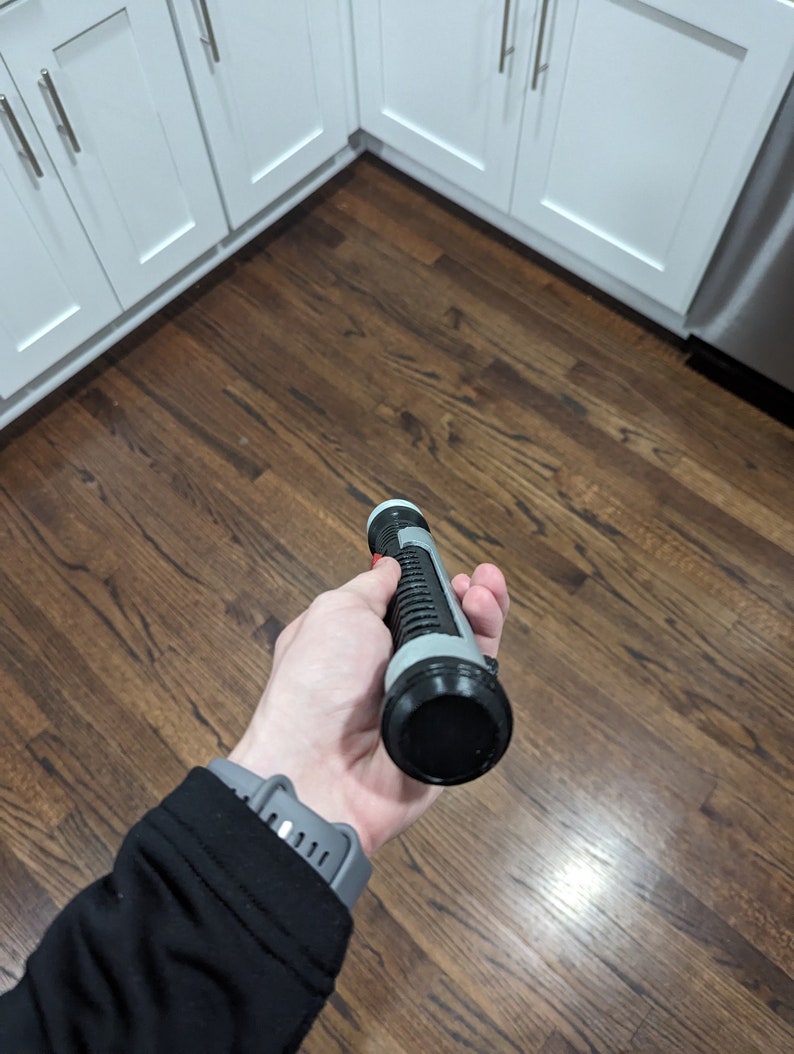 May include: A black and grey lightsaber toy with a red button. The lightsaber is being held in a hand.