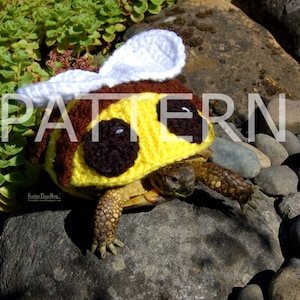 May include: A small turtle wearing a crocheted bee costume. The costume is yellow and brown with white wings and black eyes. The turtle is sitting on a rock.