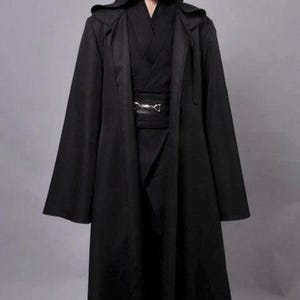 Peut inclure: Une cape à capuche noire pleine longueur portée sur une tunique croisée et un pantalon noirs. La tenue comprend une large ceinture noire fixée par une boucle en métal argenté. La cape possède de longues manches larges et une capuche profonde.