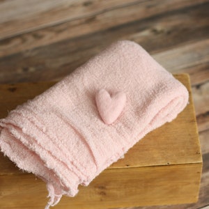 Puede incluir: Una manta rosa y esponjosa está doblada y colocada sobre una caja de madera. Un pequeño corazón de fieltro rosa está encima de la manta.