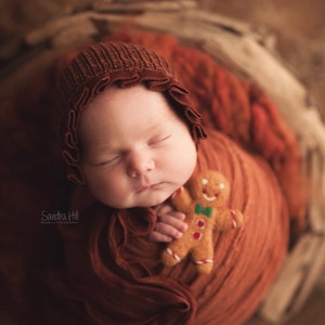 May include: A sleeping baby wrapped in a brown swaddle, wearing a brown knitted bonnet with a ruffle, holding a gingerbread man toy.